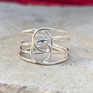 May include: A silver ring with a distinctive design. The ring has multiple bands and an open, intertwined pattern in the centre. The ring is shown on a neutral, textured surface, highlighting its intricate details.