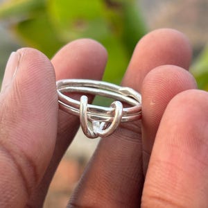 Puede incluir: Un anillo de plata con un diseño de nudo único. El anillo presenta dos bandas que se entrelazan para crear una forma de nudo. El anillo se sostiene entre los dedos, con un fondo borroso de follaje verde.