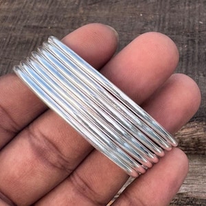 May include: A stack of silver-colored bangles, each with a rounded, ridged design. The bangles are held in a hand, with the light reflecting off their polished surfaces. The overall aesthetic is simple and elegant.