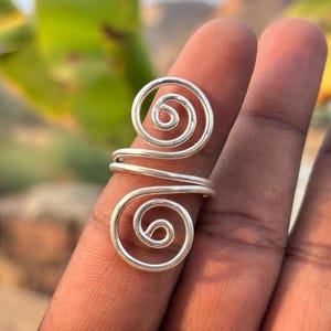 Può includere: Un anello color argento con un design a spirale. L'anello presenta due spirali vorticose collegate da una fascia. L'anello è realizzato in metallo lucido e viene mostrato su un dito.