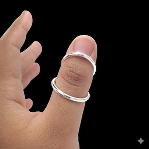 May include: Two silver rings are worn on a thumb against a black background. The rings are simple, circular bands, likely made of sterling silver or a similar metal. The rings are positioned on the thumb, one at the base of the nail and the other at the first knuckle.