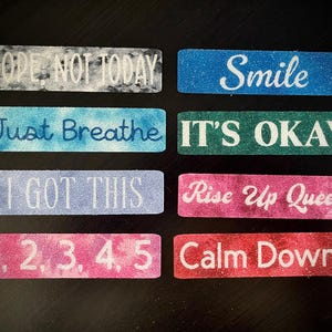 May include: Rectangular glittery stickers with motivational phrases in white. Phrases include "Nope Not Today", "Smile", "Just Breathe", "It's Okay", "I Got This", "Rise Up Queen", "1, 2, 3, 4, 5", and "Calm Down".