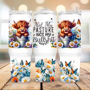 May include: White tumbler with a handle, featuring a cartoon Highland cow in a bucket of flowers. The design includes butterflies and the text "Not My Pasture Not My Bullshit". The bottom of the tumbler has a floral and butterfly design.