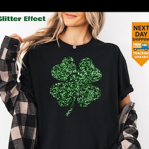 May include: Black t-shirt with a large, glittery green four-leaf clover design. The words "Glitter Effect" are above the shirt. The shirt is worn with a black and brown plaid shirt. A yellow and orange sign says "Next Day Shipping" and "Tracking Available".