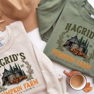 May include: Two sweatshirts, one cream and one olive green, with the text "Hagrid's Pumpkin Farm" and a graphic of a house and pumpkins. A tan hat and a fall leaf are also in the image. The sweatshirts are long-sleeved.