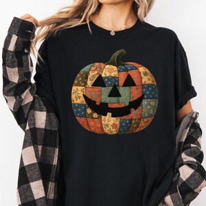 May include: Black t-shirt with a patchwork pumpkin design. The pumpkin has a smiling face and is made of various fabric patterns in orange, yellow, blue, and green. The shirt is worn with a black and beige plaid flannel.