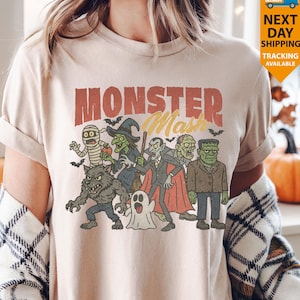 May include: A beige t-shirt with a retro graphic of Halloween monsters: a mummy, witch, vampire, and Frankenstein's monster. The words "Monster Mash" are printed above the characters. The shirt has rolled-up sleeves and a vintage aesthetic.