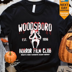 May include: Black t-shirt featuring a white and red graphic. The design includes the word "WOODSBORO" with a dripping blood effect, a ghost face, and the text "HORROR FILM CLUB". The shirt also has the text "EST. 1996" and "WHAT'S YOUR FAVORITE SCARY MOVIE?"