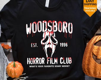Pode incluir: Camiseta preta com um design gráfico branco e vermelho. O design inclui a palavra "WOODSBORO" com efeito de sangue escorrendo, um rosto de fantasma e o texto "HORROR FILM CLUB". A camisa também tem o texto "EST. 1996" e "WHAT'S YOUR FAVORITE SCARY MOVIE?"
