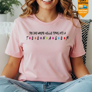 May include: A light pink t-shirt with the text "THE ONE WHERE MOLLIE TURNS INTO A TEENAGER" in black, with colorful dots between the letters. The shirt is short-sleeved and made of a soft material.