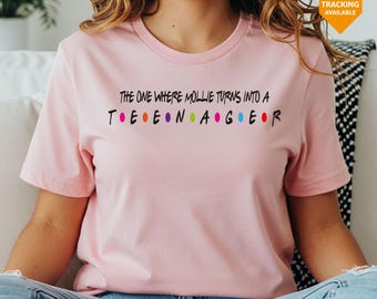 May include: A pale pink t-shirt featuring the phrase "THE ONE WHERE MOLLIE TURNS INTO A TEENAGER" in black, with coloured dots between the letters. The top is short-sleeved and made of a soft fabric.
