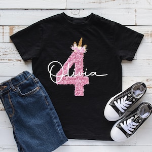 May include: Black t-shirt featuring a pink glittery number 4 unicorn design and the name "Olivia" in white script. The image includes blue denim jeans and black canvas trainers with white soles and laces.