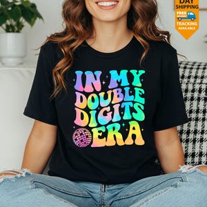 May include: Black t-shirt with the text "IN MY DOUBLE DIGITS ERA" in colorful, retro-style lettering. A disco ball graphic is below the text. The shirt is worn by a person.