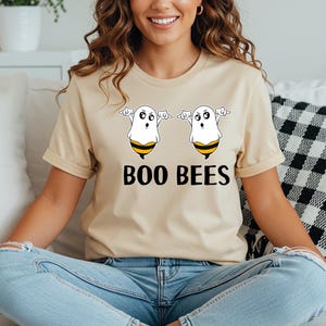 May include: Beige t-shirt featuring a Halloween-themed graphic. The design includes two cartoon ghosts with bee-like stripes and the text "BOO BEES" in black. The shirt is a casual, short-sleeved style.