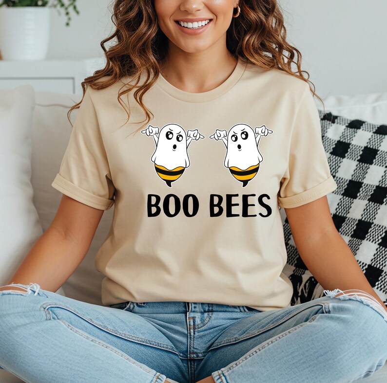 May include: Beige t-shirt featuring a Halloween-themed graphic. The design includes two cartoon ghosts with bee-like stripes and the text "BOO BEES" in black. The shirt is a casual, short-sleeved style.