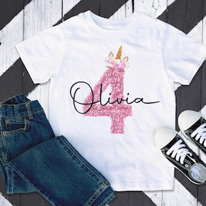 May include: White t-shirt featuring a pink glitter number 4 and the name "Olivia" in black script. The number 4 is decorated with a unicorn horn and flowers. Blue jeans and black canvas trainers are also visible.