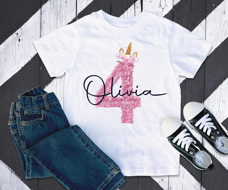 May include: White t-shirt featuring a pink glitter number 4 and the name "Olivia" in black script. The number 4 is decorated with a unicorn horn and flowers. Blue jeans and black canvas trainers are also visible.