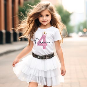 May include: A young girl in a white t-shirt featuring a pink glitter '4' and a unicorn design, with the name 'Olivia' in cursive. She wears a white tiered skirt and a belt with a silver buckle. The image is taken outdoors.