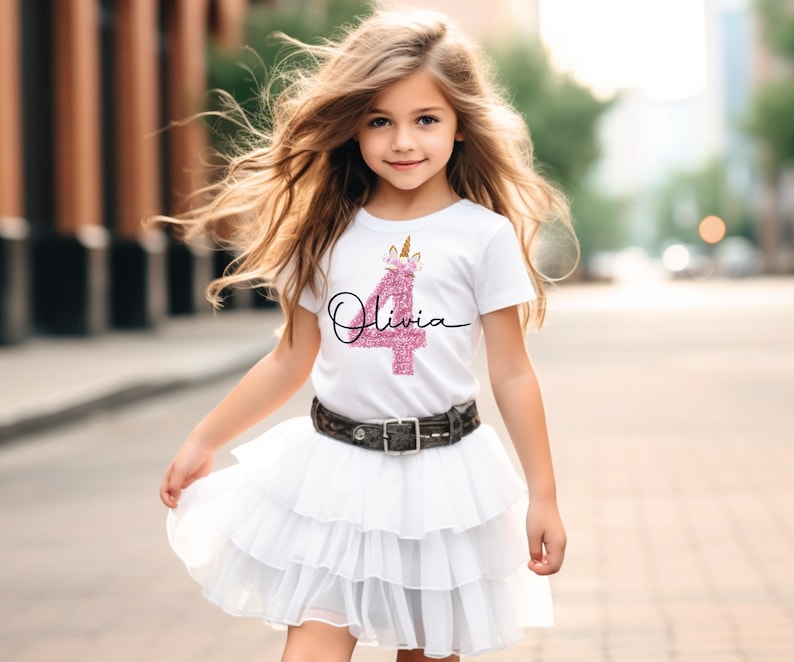 May include: A young girl in a white t-shirt featuring a pink glitter '4' and a unicorn design, with the name 'Olivia' in cursive. She wears a white tiered skirt and a belt with a silver buckle. The image is taken outdoors.