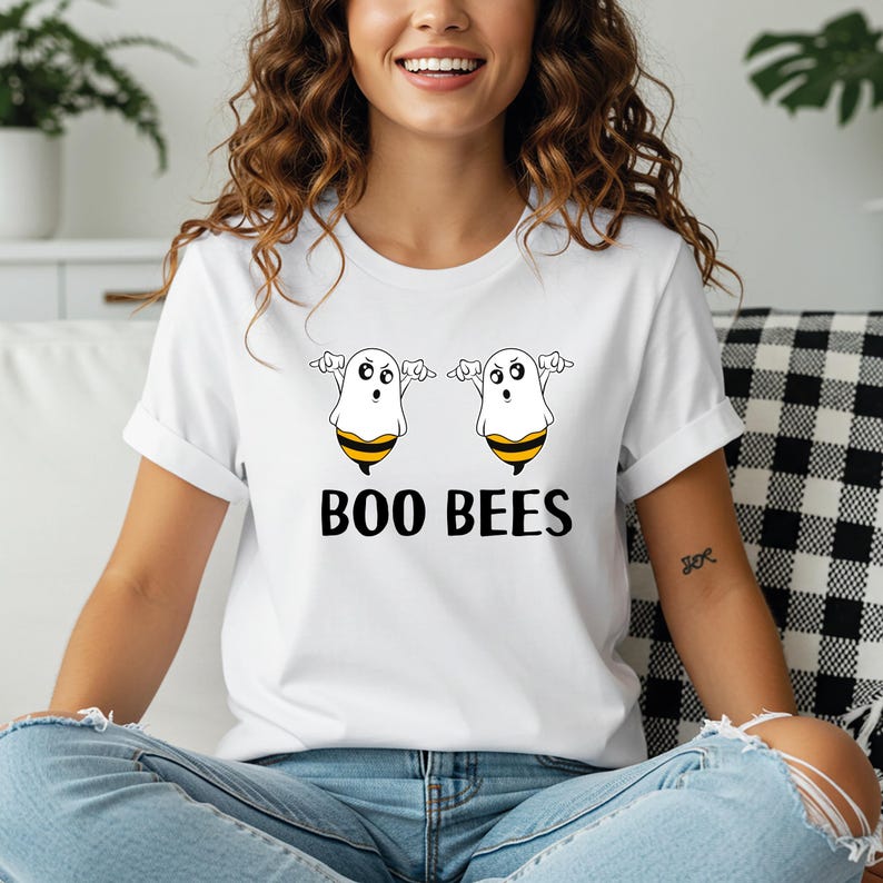 May include: White t-shirt featuring a Halloween-themed design. The shirt displays two cartoon ghost-bee hybrids with outstretched arms, and the words "BOO BEES" in bold black letters. The design is centered on the front of the shirt.