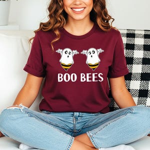 May include: Burgundy t-shirt with the text "BOO BEES" in white. The shirt features a graphic of two cartoon ghosts with bee-striped bottoms. The ghosts have outstretched arms and are smiling. The shirt is made of a soft material.