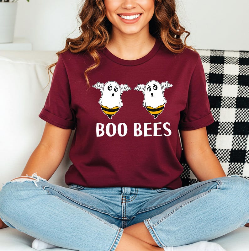 May include: Burgundy t-shirt with the text "BOO BEES" in white. The shirt features a graphic of two cartoon ghosts with bee-striped bottoms. The ghosts have outstretched arms and are smiling. The shirt is made of a soft material.