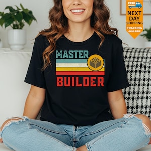 May include: Black t-shirt with a retro design featuring the words "MASTER BUILDER" in red and a brick graphic. The design includes horizontal stripes in teal, yellow, and red. A fun and playful tee for fans of building block toys.