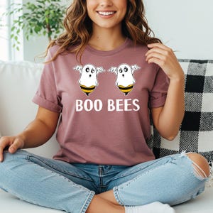 May include: A mauve t-shirt featuring the text "BOO BEES" in white. Above the text are two cartoon ghost-bee hybrids with white bodies, black eyes, and yellow and black striped bee bellies. The shirt has short sleeves and a crew neck.