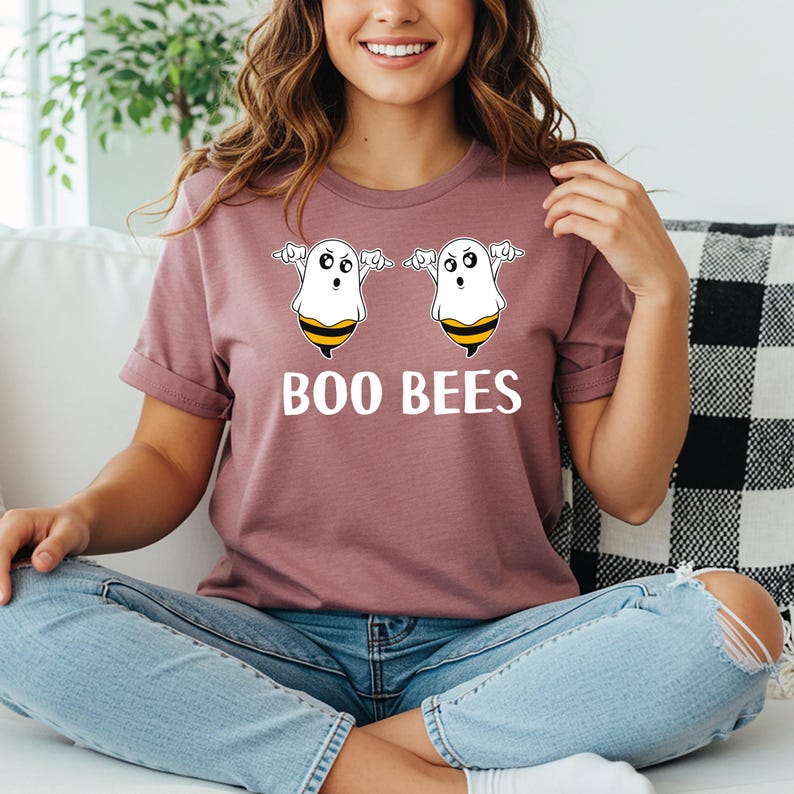 May include: A mauve t-shirt featuring the text "BOO BEES" in white. Above the text are two cartoon ghost-bee hybrids with white bodies, black eyes, and yellow and black striped bee bellies. The shirt has short sleeves and a crew neck.