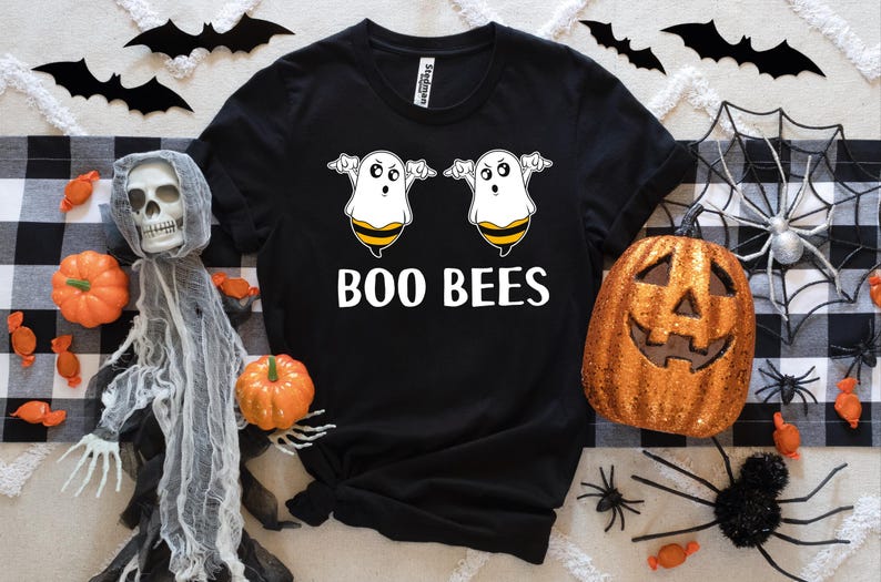 May include: Black t-shirt with the text "BOO BEES" and a graphic of two cartoon ghosts with bee stripes. The shirt is surrounded by Halloween decorations, including pumpkins, spiders, and bats.