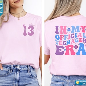 May include: Pale pink t-shirt featuring the number "13" and a crown on the front. The back displays the text "IN MY OFFICIAL TEENAGER ERA" in a retro style with floral details. A great gift for a teenager's birthday.