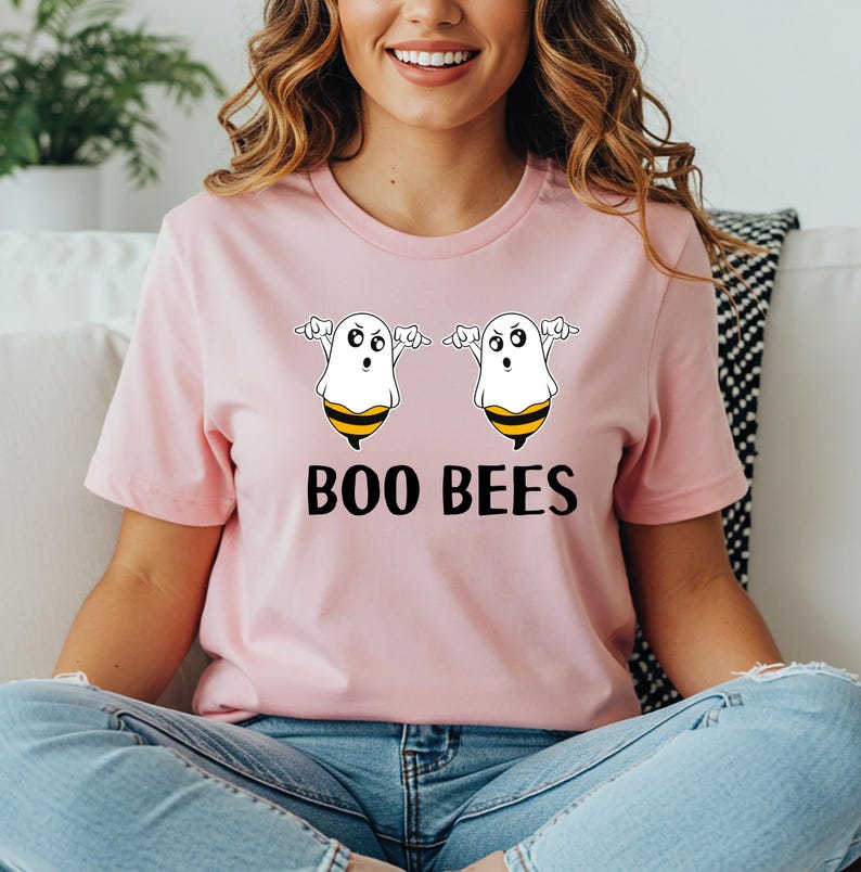May include: A light pink t-shirt featuring a graphic of two cartoon ghost-bee hybrids with the text "BOO BEES" in black. The ghost-bees have white bodies, black eyes, and yellow and black striped bee bottoms.