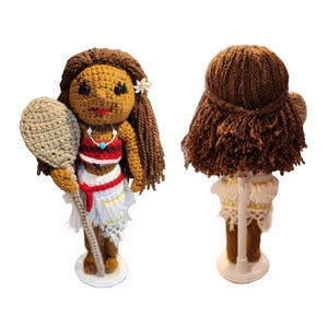 May include: A crocheted doll with brown hair, a red and white top, and a white skirt. The doll is holding a brown paddle and is standing on a white base.