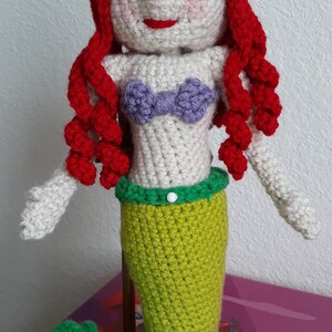 Peut inclure: Poup&eacute;e sir&egrave;ne au crochet avec des cheveux roux, un haut violet et une queue verte. La poup&eacute;e a un visage blanc avec des joues roses et des l&egrave;vres rouges. La sir&egrave;ne mesure environ 25 cm de haut.