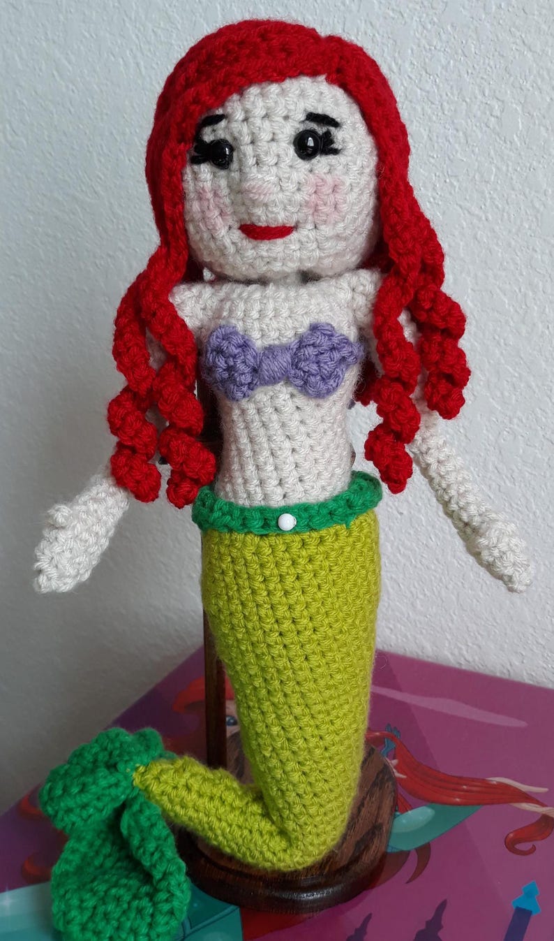 Peut inclure: Poup&eacute;e sir&egrave;ne au crochet avec des cheveux roux, un haut violet et une queue verte. La poup&eacute;e a un visage blanc avec des joues roses et des l&egrave;vres rouges. La sir&egrave;ne mesure environ 25 cm de haut.