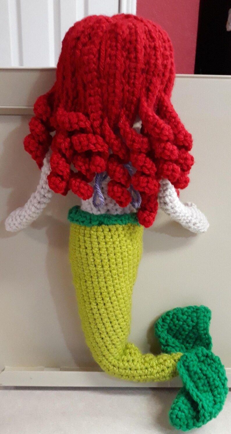 Peut inclure: Une poup&eacute;e sir&egrave;ne au crochet faite &agrave; la main avec des cheveux rouge vif, des bras blancs, une queue verte et jaune et une nageoire verte. La poup&eacute;e est faite avec du fil et a une apparence fantaisiste et artisanale. La sir&egrave;ne est un jouet doux unique.