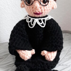 May include: Crocheted doll of a woman wearing a black robe, white collar, and glasses. The doll has brown hair and a pink face.