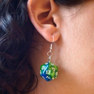 May include: A single earring featuring a blue and green D20 die, suspended from a silver hook. The die has white numbers and is worn on the ear. The earring is a unique accessory.