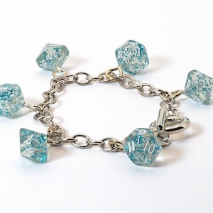 May include: A silver-toned chain bracelet featuring six clear, blue-glitter dice charms. The dice are of various shapes, with white numbering. The bracelet has a toggle clasp and is set against a white background.