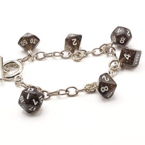 May include: A silver chain bracelet featuring six dark brown, speckled dice charms. Each die has white numbers. The bracelet has a toggle clasp. The dice are in various positions, displaying different numbers.