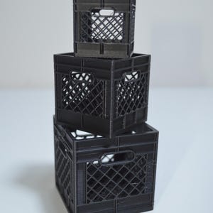 May include: Three black plastic milk crates stacked on top of each other. The crates are made of a grid pattern and have a handle on the front.