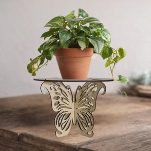 May include: A decorative wooden butterfly plant stand with a round top, holding a terracotta pot with a green plant. The butterfly design is intricately cut out of the wood, creating an airy and elegant look. The plant has green and white leaves.