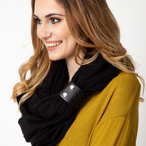 May include: A black infinity scarf with a black leather strap with silver snaps. The scarf is worn by a person with long brown hair and a yellow shirt.