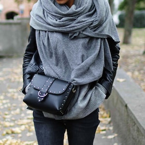 May include: A woman wearing a gray knitted scarf, a black leather jacket, and dark blue jeans. She is carrying a black leather satchel with silver studs and a silver buckle.