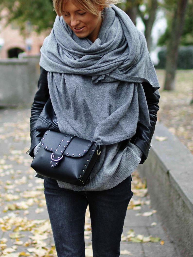 May include: A woman wearing a gray knitted scarf, a black leather jacket, and dark blue jeans. She is carrying a black leather satchel with silver studs and a silver buckle.