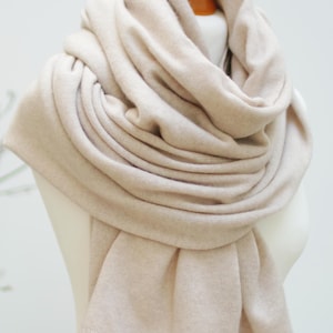 Può includere: Una sciarpa in maglia color beige drappeggiata in più strati attorno a un manichino. La sciarpa presenta una consistenza fine e liscia con una tonalità marrone chiaro uniforme, caratterizzata da un design semplice e pulito con bordi rifiniti.