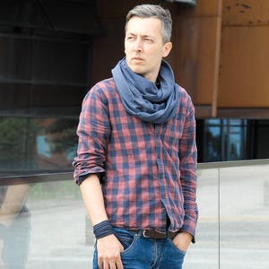 Puede incluir: Un hombre con una camisa de cuadros rojos y negros con una bufanda azul alrededor del cuello. Está de pie frente a una barandilla de vidrio y mirando hacia un lado.