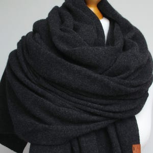 May include: A charcoal grey, oversized scarf draped over a mannequin. The scarf is made of a thick, soft material and is wrapped around the neck and shoulders. A small, brown leather tag is visible on the edge. The scarf appears to be warm and cosy.