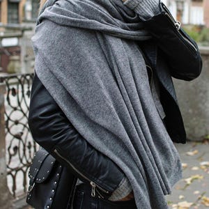 May include: A woman wearing a gray scarf, a black leather jacket, and dark denim jeans. The scarf is draped over her shoulders and around her neck.