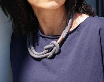 Puede incluir: Un collar de cuerda gris con un acento metálico. El collar está anudado y tiene un diseño simple.